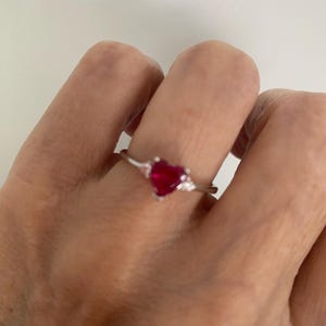 May include: A silver-toned ring featuring a central heart-shaped red gemstone flanked by two smaller, clear accent stones on either side of the band. The ring has a polished metal finish and a simple, slender band design that sits on the finger.