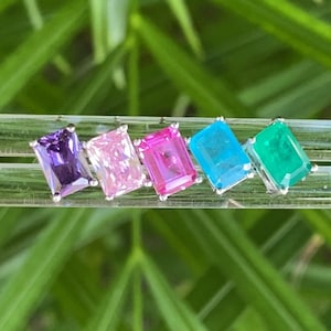 May include: Five emerald-cut gemstones in a rainbow of colors: purple, pink, hot pink, blue, and green. The gemstones are set in silver settings and are arranged in a row.
