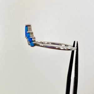 May include: A silver ring with a row of blue opal stones. The ring is held by a pair of black tweezers against a white background. The ring's band is thin and polished.