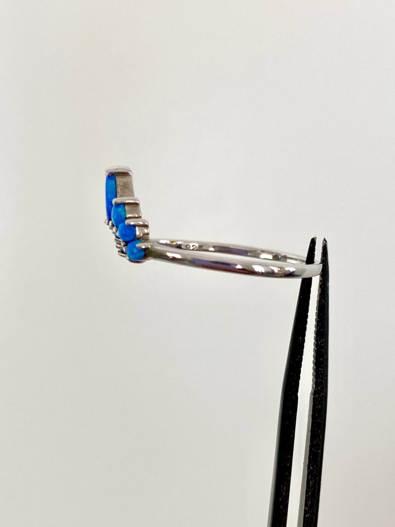 May include: A silver ring with a row of blue opal stones. The ring is held by a pair of black tweezers against a white background. The ring's band is thin and polished.