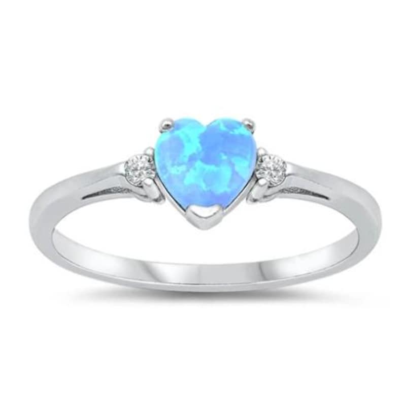 May include: A silver ring featuring a heart-shaped, light blue opal center stone. Two small, clear accent stones flank the opal. The ring has a polished, shiny finish, and a simple band.