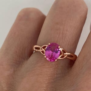 May include: Rose gold ring featuring a large, oval-cut pink gemstone. The ring has a Celtic knot design on the band, adding a touch of elegance. The gemstone is the focal point, showcasing a vibrant pink hue.