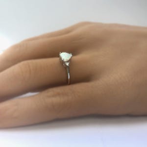 May include: A silver ring with a pear-shaped opal gemstone and two smaller accent stones. The ring is set on a hand with pink nail polish.