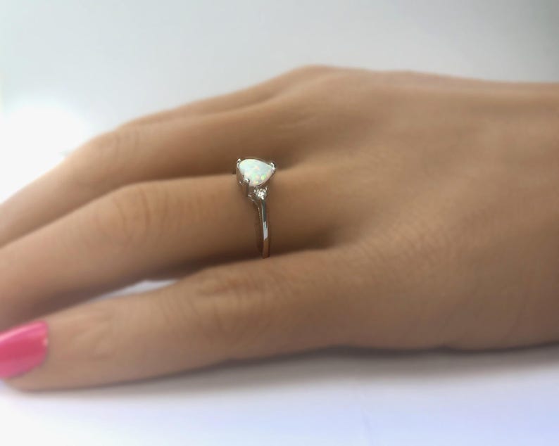 May include: A silver ring with a pear-shaped opal gemstone and two smaller accent stones. The ring is set on a hand with pink nail polish.
