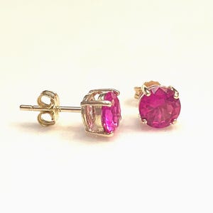 May include: A pair of stud earrings featuring bright pink gemstones set in gold-colored metal. The gemstones are round and faceted, reflecting light. The earrings are displayed on a white background.