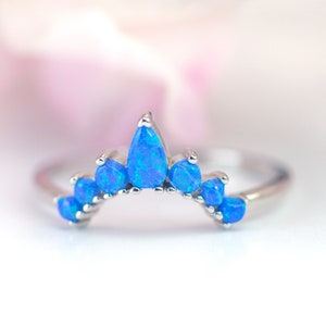 May include: A silver ring featuring a crown design with vibrant blue opal stones. The ring's band is thin, and the opal stones are teardrop and round shaped. The ring is set against a soft, blurred background.