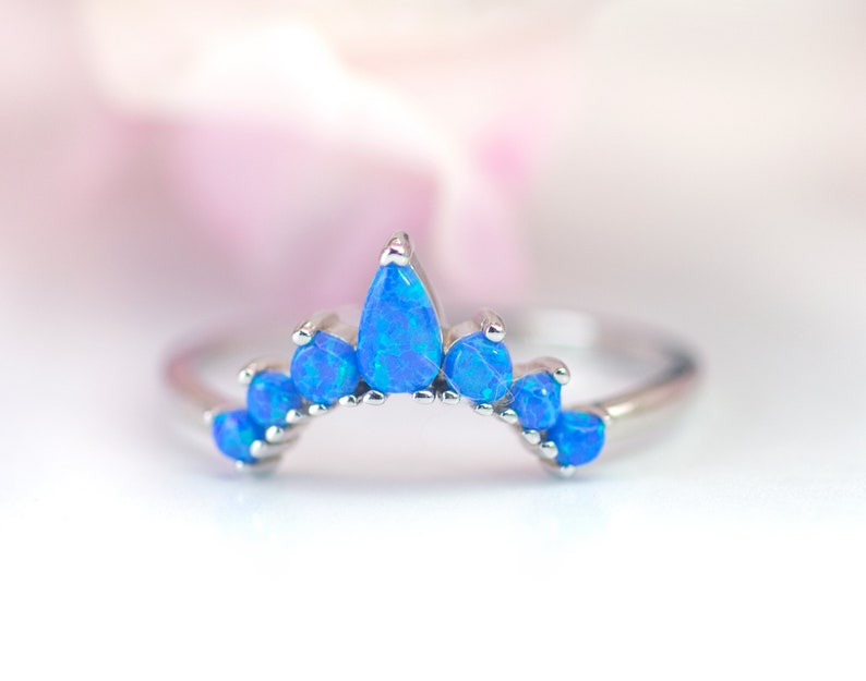 May include: A silver ring featuring a crown design with vibrant blue opal stones. The ring's band is thin, and the opal stones are teardrop and round shaped. The ring is set against a soft, blurred background.