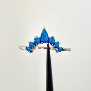 May include: A silver ring featuring a curved design with multiple blue opal stones. The ring has a crown-like shape, with a larger teardrop-shaped stone at the center and smaller round stones on either side. The stones have a vibrant blue color.