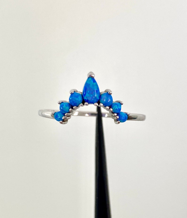 May include: A silver ring featuring a curved design with multiple blue opal stones. The ring has a crown-like shape, with a larger teardrop-shaped stone at the center and smaller round stones on either side. The stones have a vibrant blue color.