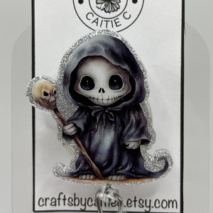 May include: A badge reel featuring a cartoon grim reaper character with a skull staff. The reaper is wearing a black hooded cloak and has a silver glitter border. The text "Crafts by Caitie C" and "craftsbycaitie.etsy.com" are also visible.