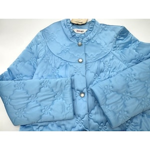 May include: Light blue quilted jacket with a floral pattern. The jacket features a round collar, button closure, and long sleeves. The fabric has a subtle sheen, and the floral design is stitched throughout the garment.