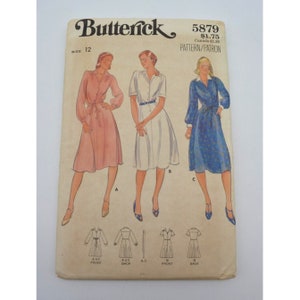 May include: Vintage sewing pattern for a dress, Butterick 5879, size 12. The pattern shows three different styles of dresses: a pink dress with long sleeves, a white dress with short sleeves, and a blue floral dress with long sleeves. The pattern includes instructions for sewing the dress.