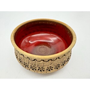 May include: A round ceramic bowl with a textured, light brown exterior and a glossy red interior. The exterior features a repeating floral pattern. The rim is a lighter shade of brown. The bowl is likely for decorative or serving purposes.