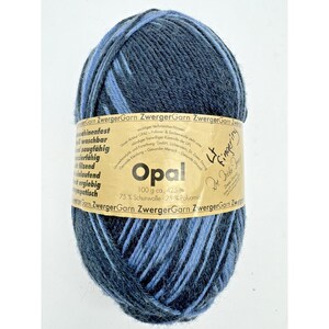 May include: A ball of variegated yarn in shades of dark blue and light blue. A tan paper label wraps around the center, featuring the text Opal, 100 g ca. 425 m, 75% Schurwolle, 25% Polyamid, and ZwergerGarn, along with additional German text.
