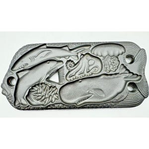 May include: A rectangular cast iron mould featuring embossed sea creatures including a shark, dolphin, anemone, octopus, jellyfish, and whale. Each figure is labelled with its name in raised text. The mould has a textured border and three holes.
