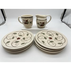May include: A ceramic tea set featuring a cream-colored mug, a cream-colored creamer pitcher, and six matching plates. Each piece is decorated with a brown swirling vine pattern, green leaves, and a small red flower motif in the center.