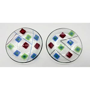 May include: Two clear glass plates with a black rim. Each plate features a design of red, blue, and green square shapes. Thin lines of color and small dots add to the decorative pattern. A unique addition to any table setting.