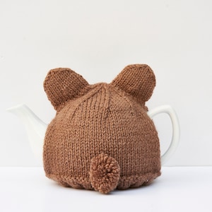Puede incluir: Calienta-tetera de punto marrón con orejas y cola de oso. El calienta-tetera está en una tetera blanca.
