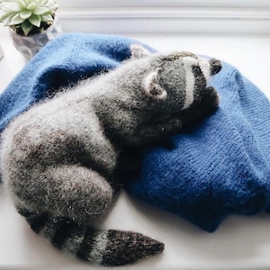 Könnte beinhalten: Ein handgefertigtes graues und weißes Waschbär-Plüschtier, das auf einem blauen Pullover liegt. Der Waschbär hat einen gestreiften Schwanz und einen weißen Streifen über dem Gesicht. Eine kleine Sukkulentenpflanze in einem grauen Topf befindet sich im Hintergrund.