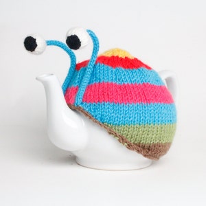 Puede incluir: Una tetera blanca con un gorro de punto de rayas de colores y ojos saltones. El gorro tiene rayas rojas, rosas, azules, verdes y marrones.