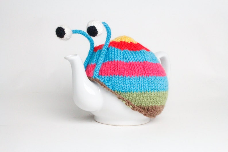 Puede incluir: Una tetera blanca con un gorro de punto de rayas de colores y ojos saltones. El gorro tiene rayas rojas, rosas, azules, verdes y marrones.