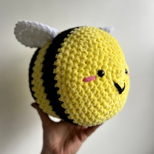 Pode incluir: Um brinquedo de abelha de pelúcia crochetado com listras amarelas e pretas, asas brancas e um rosto sorridente. A abelha tem bochechas cor-de-rosa e olhos pretos.