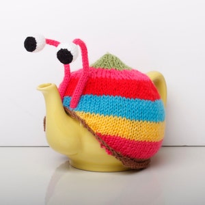 以下が含まれることがあります： 黄色いティーポットに、カラフルなストライプのニットのカバーと、ピンクと白のググリーアイが2つ付いている。