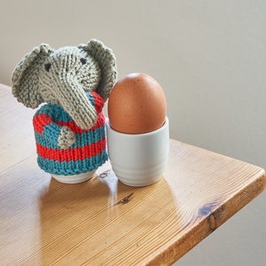 May include: A knitted gray elephant wearing a blue and red striped sweater sits next to a brown egg in a white egg cup on a wooden table.
