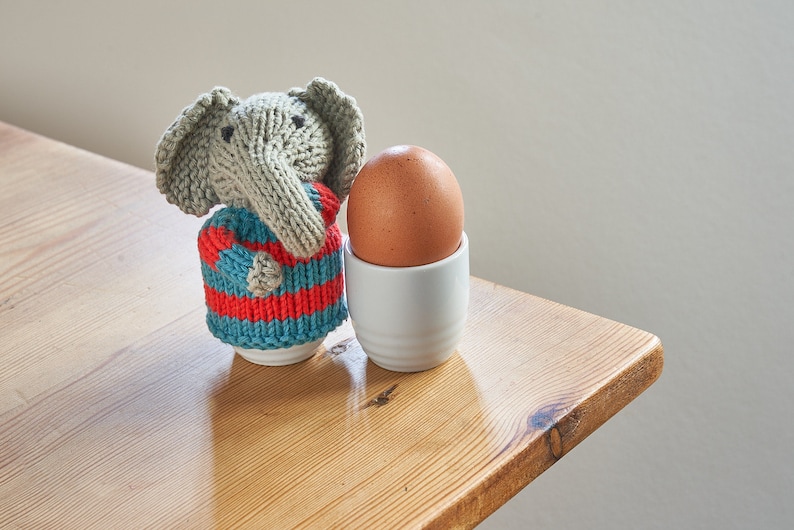 May include: A knitted gray elephant wearing a blue and red striped sweater sits next to a brown egg in a white egg cup on a wooden table.