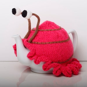 Puede incluir: Una funda de tetera de punto rosa con detalles marrones y grandes ojos blancos. La funda tiene forma de caracol y está sobre una tetera blanca.