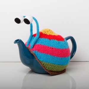 Puede incluir: Una tetera azul con un calienta-tetera de punto de colores con forma de caracol con ojos saltones.