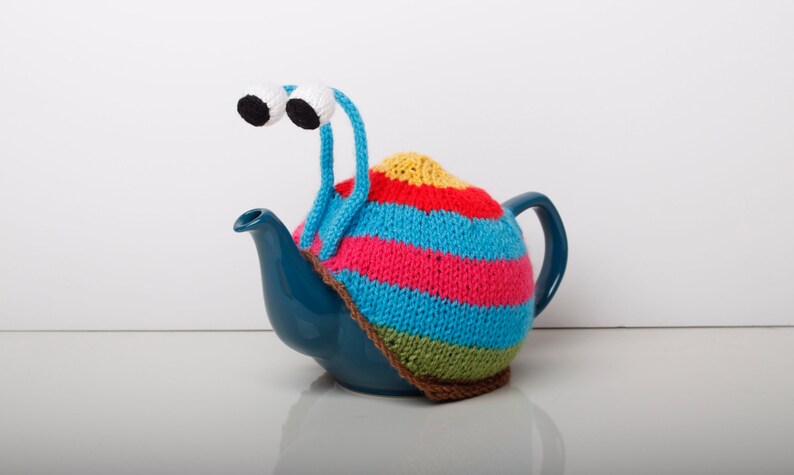Puede incluir: Una tetera azul con un calienta-tetera de punto de colores con forma de caracol con ojos saltones.