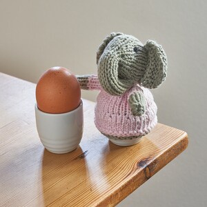 May include: A knitted elephant toy wearing a pink dress, standing next to a brown egg in a white egg cup on a wooden table.
