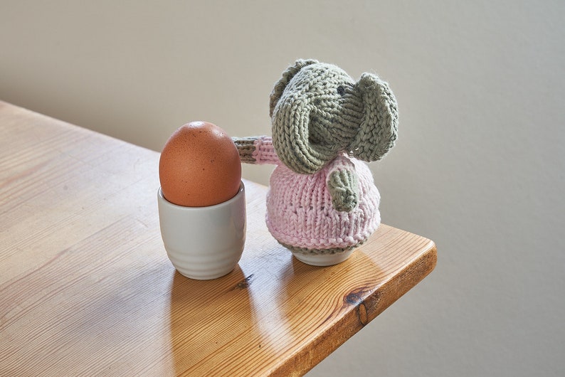 May include: A knitted elephant toy wearing a pink dress, standing next to a brown egg in a white egg cup on a wooden table.
