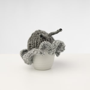 May include: A gray knitted egg cozy with two black eyes and a long gray stem. The cozy is sitting on top of a white egg.