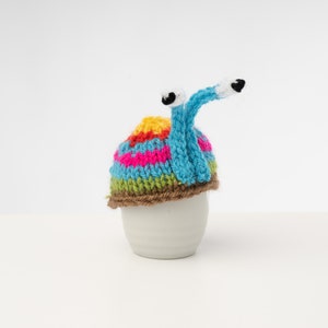 Puede incluir: Un gorro de ganchillo de caracol colorido con rayas marrones, azules, verdes, rosas y amarillas. El gorro tiene dos ojos blancos con pupilas negras y una larga cola azul de ganchillo.