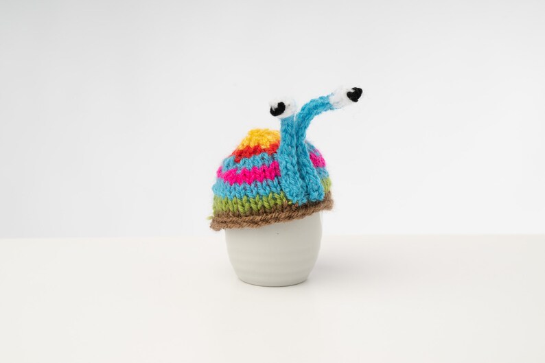 Puede incluir: Un gorro de ganchillo de caracol colorido con rayas marrones, azules, verdes, rosas y amarillas. El gorro tiene dos ojos blancos con pupilas negras y una larga cola azul de ganchillo.