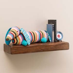 May include: A colorful crocheted dachshund dog toy, lying on a wooden shelf with books and a clock. The dog is striped with brown, orange, yellow, pink, and blue, with a turquoise blue nose and ears.