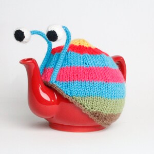 Puede incluir: Una tetera roja con una cubierta de punto de rayas de colores y ojos saltones. La cubierta tiene rayas azules, rosas, amarillas, verdes y marrones.