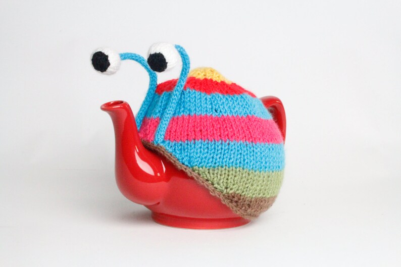 Puede incluir: Una tetera roja con una cubierta de punto de rayas de colores y ojos saltones. La cubierta tiene rayas azules, rosas, amarillas, verdes y marrones.