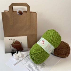 Könnte beinhalten: Ein Häkelset mit grünem und braunem Garn, einem weißen Pompon, einer Häkelnadel und Anleitungen zum Häkeln einer Schnecke. Das Set wird in einer braunen Papiertüte geliefert.