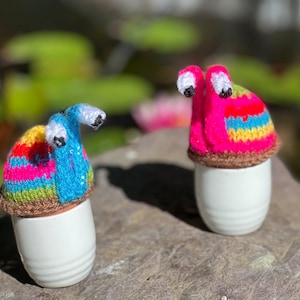 Puede incluir: Dos figuritas de caracol de crochet de colores, una rosa y otra azul, con ojos blancos, sentadas sobre recipientes de cer&aacute;mica blancos en forma de huevo.