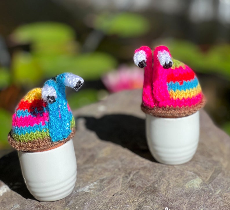 Puede incluir: Dos figuritas de caracol de crochet de colores, una rosa y otra azul, con ojos blancos, sentadas sobre recipientes de cer&aacute;mica blancos en forma de huevo.