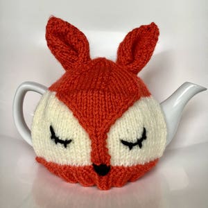 Puede incluir: Un calienta-tetera de punto naranja y blanco con forma de zorro con los ojos cerrados y una nariz negra.