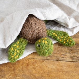 Pode incluir: Um brinquedo de tartaruga de malha marrom e verde espiando por baixo de um cobertor de linho branco. A tartaruga tem detalhes amarelos em sua carapaça e membros.