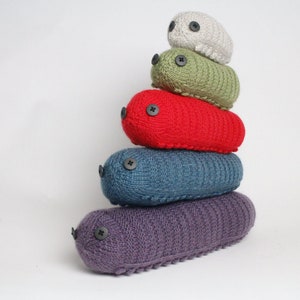 May include: A stack of five knitted, oblong-shaped creatures in various colors: purple, teal, red, green, and gray. Each has a button eye. The creatures are stacked on top of each other against a white background.