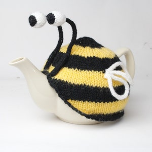 以下が含まれることがあります： 白いティーポットに、黄色と黒のニットでできた蜂のティーコージーがかけられています。コージーには白い目と触角が付いています。