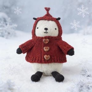 Peut inclure: Une peluche tricotée à la main dans un paysage enneigé. Le jouet en peluche est blanc avec une veste à capuche rouge fermée par des boutons en bois en forme de cœur. Il a des pattes et des oreilles noires, et une petite touffe sur la capuche.