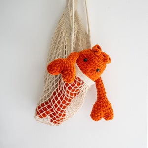 Könnte beinhalten: Ein gehäkeltes orangefarbenes Hummer-Spielzeug, das in einer weißen Netztasche hängt. Der Hummer hat schwarze Augen und eine strukturierte Oberfläche.