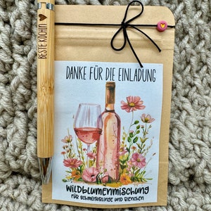 Könnte beinhalten: Eine braune Papiertüte mit einem rosa Herzaufkleber und einem schwarzen Band, das um die Oberseite gebunden ist. Die Tüte hat ein weißes Etikett mit dem Text "Danke für die Einladung" und einer Aquarell-Illustration eines Glases rosa Wein und einer Flasche rosa Wein mit rosa Blumen. Das Etikett trägt auch den Text "Wildblumenmischung für Schmetterlinge und Bienen".